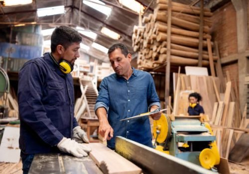 Latin American foreman talking to a worker cutting planks of wood at a timber factory